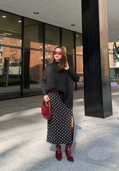 Mujer con top negro, falda de lunares negros y blancos, botas rojas y gafas de sol, sosteniendo un bolso rojo frente a un edificio moderno.