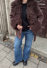 Vêtement d'extérieur marron avec un col et des poignets en fourrure, porté sur un pull noir, associé à un jean bleu à jambes larges et des bottines noires.