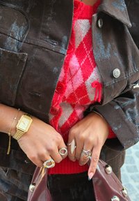 Veste marron à motif subtil, pull rouge à losanges, plusieurs bagues, montre en or, sac bordeaux, boutons texturés. Accents de bijoux remarquables.