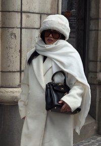 Femme portant un chapeau en fourrure blanche, un manteau blanc et une écharpe, tenant un sac à main noir, debout près de l'entrée d'un bâtiment en pierre.