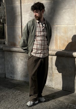 Hombre con cabello rizado y barba, vestido con camisa de cuadros, chaqueta verde y pantalones marrones, de pie contra una pared de piedra con las manos detrás de la espalda.