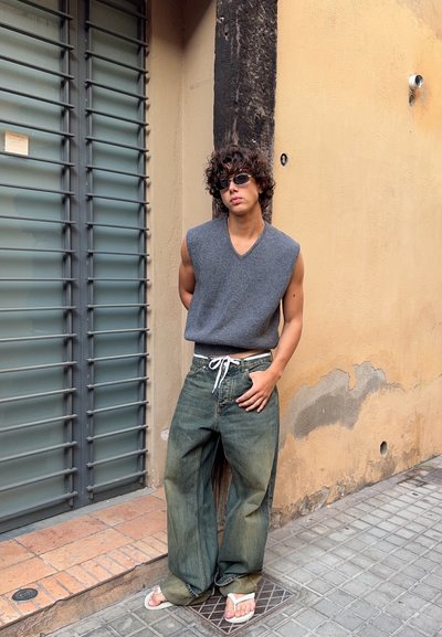 Joven con cabello rizado que lleva gafas de sol, top sin mangas gris, vaqueros holgados y chanclas blancas apoyado contra una pared beige cerca de una persiana metálica.