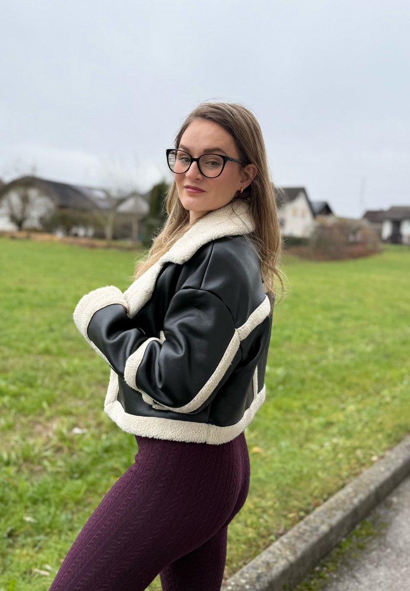 Black leather jacket with white shearling collar and trim, paired with purple textured leggings, captured outdoors with a grassy background.