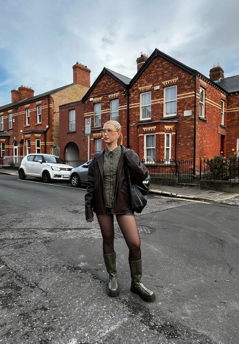 Young woman wearing black jacket, plaid shirt, tights, and green boots stands on a paved street with brick houses and parked cars in the background.