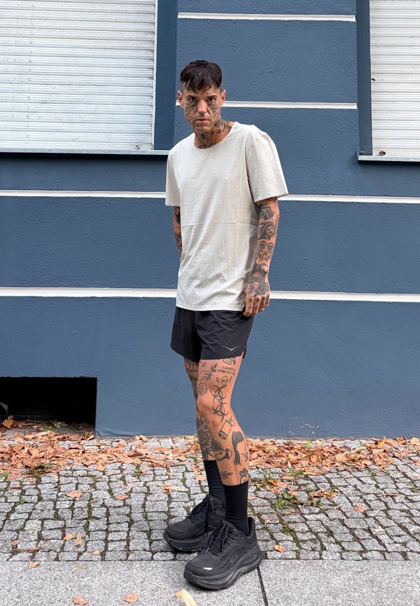 Joven con tatuajes en la cara, los brazos y las piernas, de pie sobre una acera de adoquines cubierta de hojas, frente a una pared azul grisácea con franjas blancas.