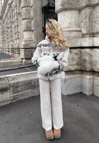 Valge karvane lukuga jakk, millel on põhjapõdra muster, kombineeritud soonikkoeliste valgete pükstega ja helepruunide platvormsaabastega, kivise taustaga.