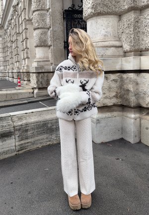 Chaqueta blanca con cremallera y diseño de renos, combinada con pantalones blancos acanalados y botas de plataforma marrón claro, sobre un fondo de piedra.