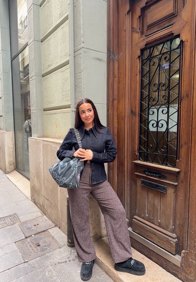 Joven con cabello largo apoyada en una pared de piedra junto a una gran puerta de madera, vistiendo una chaqueta negra, pantalones estampados y sosteniendo una bolsa grande gris.