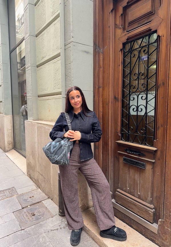 Ung kvinde med langt hår læner sig mod en stenmur ved en stor trædør, iført en sort jakke, mønstrede bukser og holder en stor grå taske.