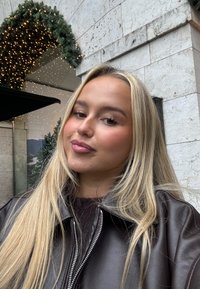 Brązowa skórzana kurtka z zamkiem z przodu, noszona na ciemnym swetrze. Długie, proste, blond włosy. Miękkie tło z migoczącymi światełkami.