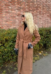 A woman with long blonde hair is wearing a brown belted coat, black gloves with fur cuffs, and sunglasses while standing next to green bushes and a brick wall.