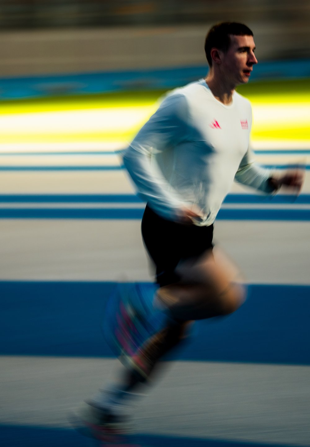 Athlète masculin portant un maillot blanc à manches longues et un short noir, courant sur une piste avec des marquages de couloir bleus et jaunes, en mouvement flou.