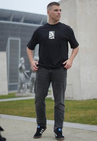 T-shirt noir à manches courtes avec un imprimé graphique au centre, associé à un jean gris foncé et des baskets noires avec des accents bleus.