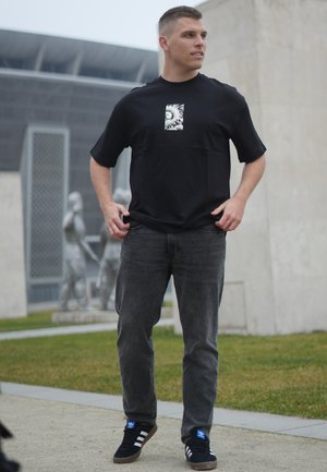 Camiseta negra de manga corta con un estampado gráfico en el centro, combinada con jeans gris oscuro y zapatillas negras con detalles en azul.