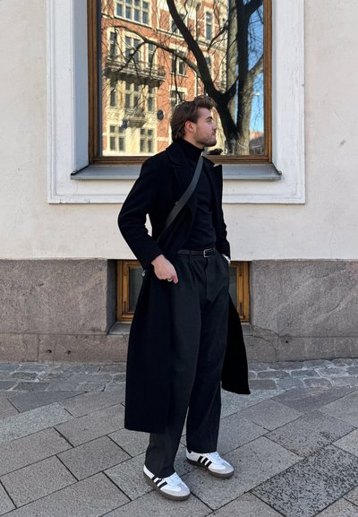 Hombre con abrigo negro, cuello de tortuga y pantalones amplios, con zapatillas blancas, está de pie sobre un pavimento de piedra, con una ventana del edificio reflejando un árbol y la arquitectura cercana.