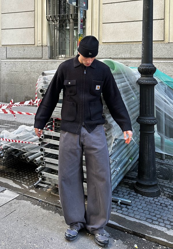 Homme portant un bonnet noir, un gilet zippé noir et un pantalon large gris, debout sur un trottoir près de tuyaux métalliques empilés et d'un lampadaire noir.