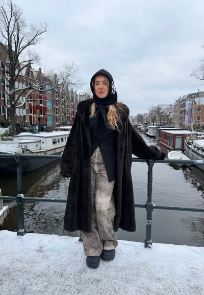 Mujer con un abrigo de piel negro y bufanda con capucha de pie en un puente sobre un canal nevado, con barcos y edificios históricos al fondo.