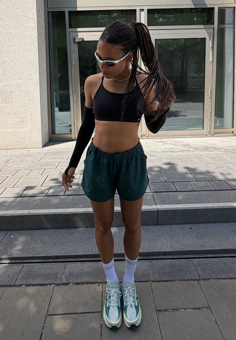 Femme aux longs cheveux tressés, portant des lunettes de soleil, un crop top noir, des manches noires, un short foncé, des chaussettes blanches et des chaussures de course vertes, debout dehors.
