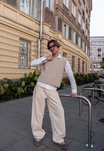 Chaleco de punto beige sobre una camisa blanca, combinado con pantalones holgados color crema. El sujeto está de pie junto a una barandilla metálica al aire libre, con vegetación.