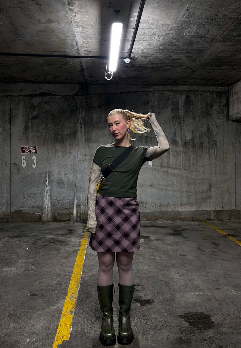 Giovane donna con coda di cavallo bionda si trova in un parcheggio sotterraneo di cemento poco illuminato, indossa una maglietta verde, gonna con motivo, maniche lunghe e stivali scuri.