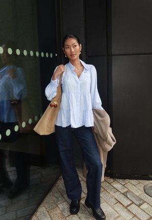 Camisa de botones azul claro con mangas largas, diseño en capas, combinada con jeans azul oscuro y un bolso beige, que presenta acentos rojos redondos.