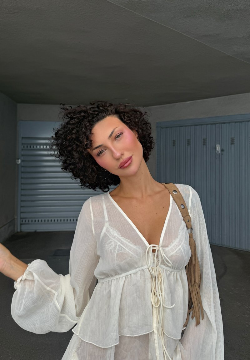 Leggera camicia bianca con maniche lunghe e puff e un orlo a strati. Presenta un fronte con cordoncino e dettagli in pizzo, abbinata a una tracolla beige.
