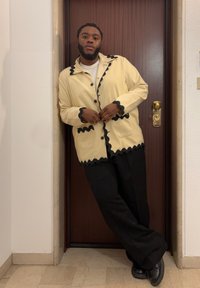 Bărbat cu barbă sprijinindu-se de o ușă din lemn, îmbrăcat într-o jachetă crem cu detalii negre, pantaloni negri și pantofi negri, într-un spațiu închis.