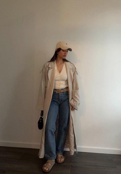 Gabardina beige sobre una camiseta blanca de canalé, combinada con jeans azules y sandalias beige. Los accesorios incluyen una gorra de color claro y un bolso negro.