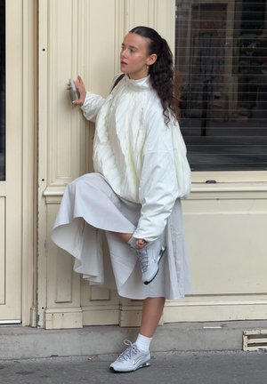 Chaqueta acolchada blanca con pliegues, falda de color gris claro hasta las rodillas y zapatillas grises. El sujeto ajusta su zapato mientras se apoya en una puerta.