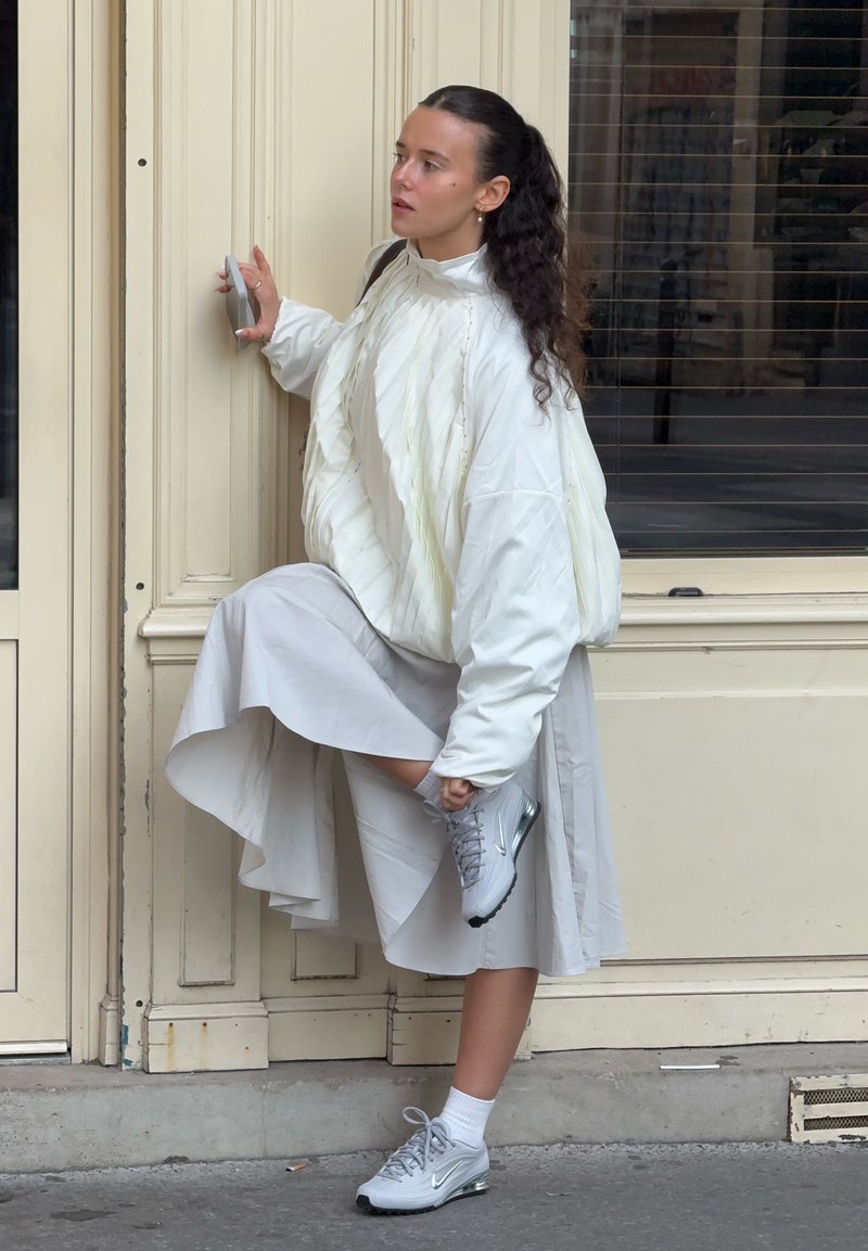 White puffer jacket with pleats, knee-length light grey skirt, and grey sneakers. Subject adjusts shoe while leaning against a door.
