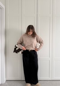 Femme ajustant un pull beige rentré dans un pantalon large noir, tenant un sac à main noir et blanc à motifs, debout devant des portes blanches à panneaux.
