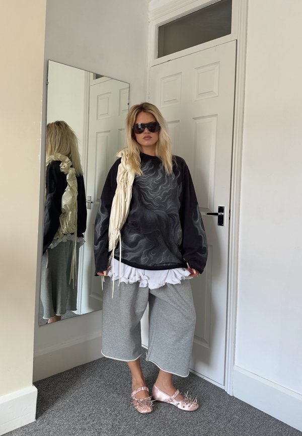 Mujer con suéter negro de gran tamaño, pantalones anchos grises, camisa blanca con volantes, grandes gafas de sol y zapatos rosas con picos, de pie dentro de una habitación cerca de un espejo.