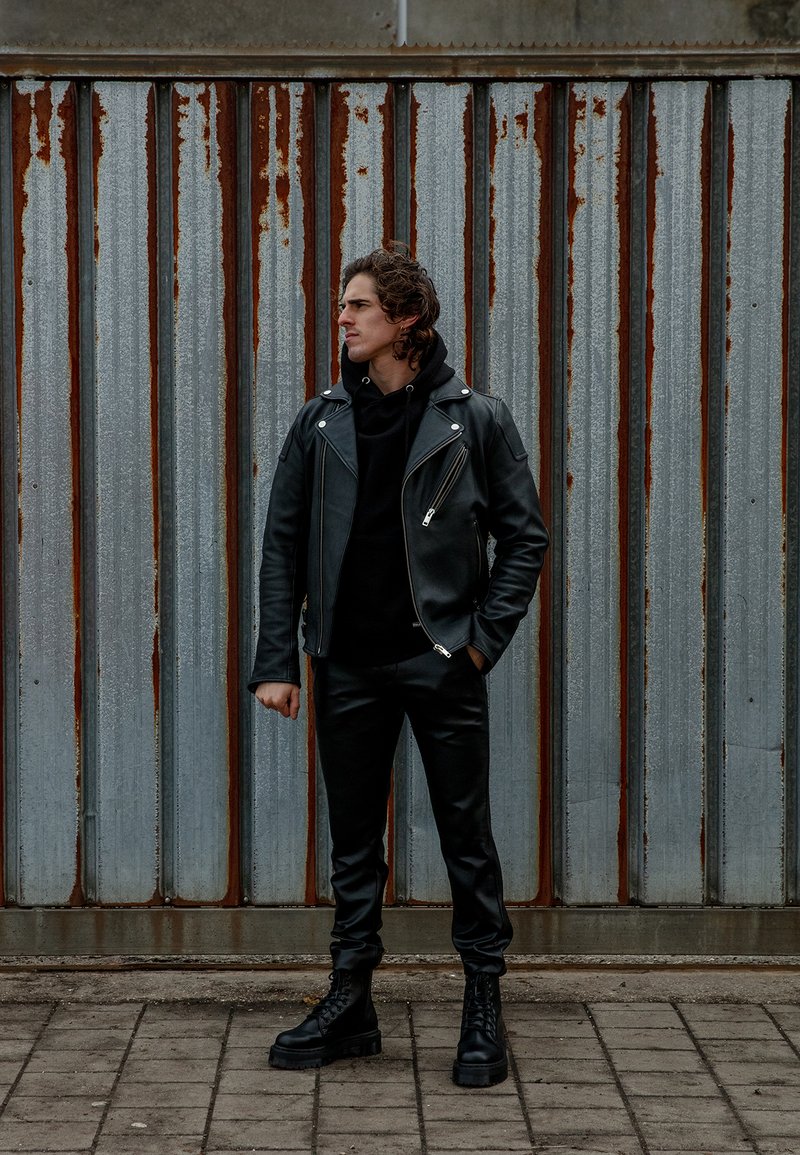 Juodas odinis motociklininkų švarkas su sidabrinėmis detalėmis, juoda džemperis, prigludę juodi kelnės ir masyvūs juodi batai prieš gofruotą metalinę fono.