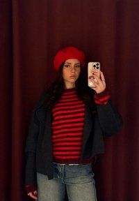 Béret rouge, pull à rayures rouges et noires, veste grise et jean bleu. Texture lisse, coupe décontractée, avec des étiquettes visibles sur la coque du téléphone.