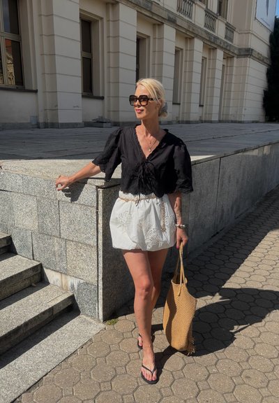 Blusa negra de manga corta con encaje y una mini falda blanca bordada. Lleva un bolso de mano tejido en color marrón claro y chanclas negras.