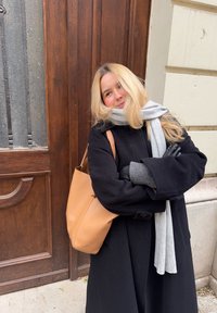 Jeune femme aux cheveux blonds portant un manteau noir, une écharpe grise et des gants, debout souriante, les bras croisés près d'une porte en bois, avec un sac à bandoulière en cuir beige.