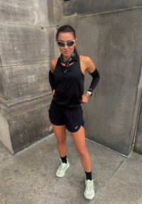 Débardeur sportif noir avec découpes, shorts noirs, manchons de bras noirs, bandana noir et blanc à motifs, et baskets vert clair.
