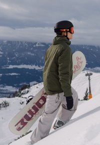Snøbrettkjører som klatrer opp en snødekt bakke, iført en olivengrønn jakke, brune bukser, svarte hansker og en svart hjelm med oransje detaljer, mens han holder et hvitt snøbrett.