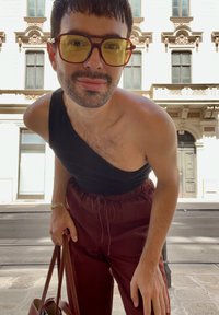 Top senza maniche nero, pantaloni bordeaux, grandi occhiali giallo-tinti, con una borsa marrone, in piedi all'aperto con uno sfondo di edificio ornato.