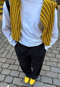 Wit overhemd met lange mouwen, zwarte loszittende broek en gele sneakers. Over de schouders hangt een geel gebreide trui met gestructureerde details.