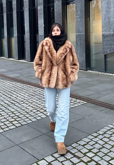 Abrigo de piel sintética marrón con un acabado texturizado, combinado con jeans de color azul claro y botas de plataforma color tostado. Una bufanda negra completa el conjunto.