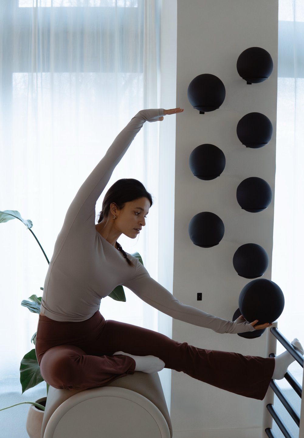 Femme en tenue de sport étirant une jambe sur une barre tout en s'équilibrant sur un équipement d'exercice arrondi dans une pièce lumineuse avec des sphères noires sur le mur.