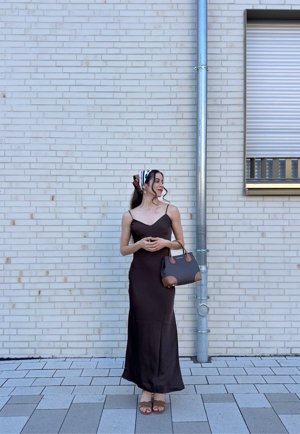 Femme en longue robe sombre et foulard sur la tête, debout sur un trottoir carrelé contre un mur de briques claires, tenant un sac à main et regardant de côté.