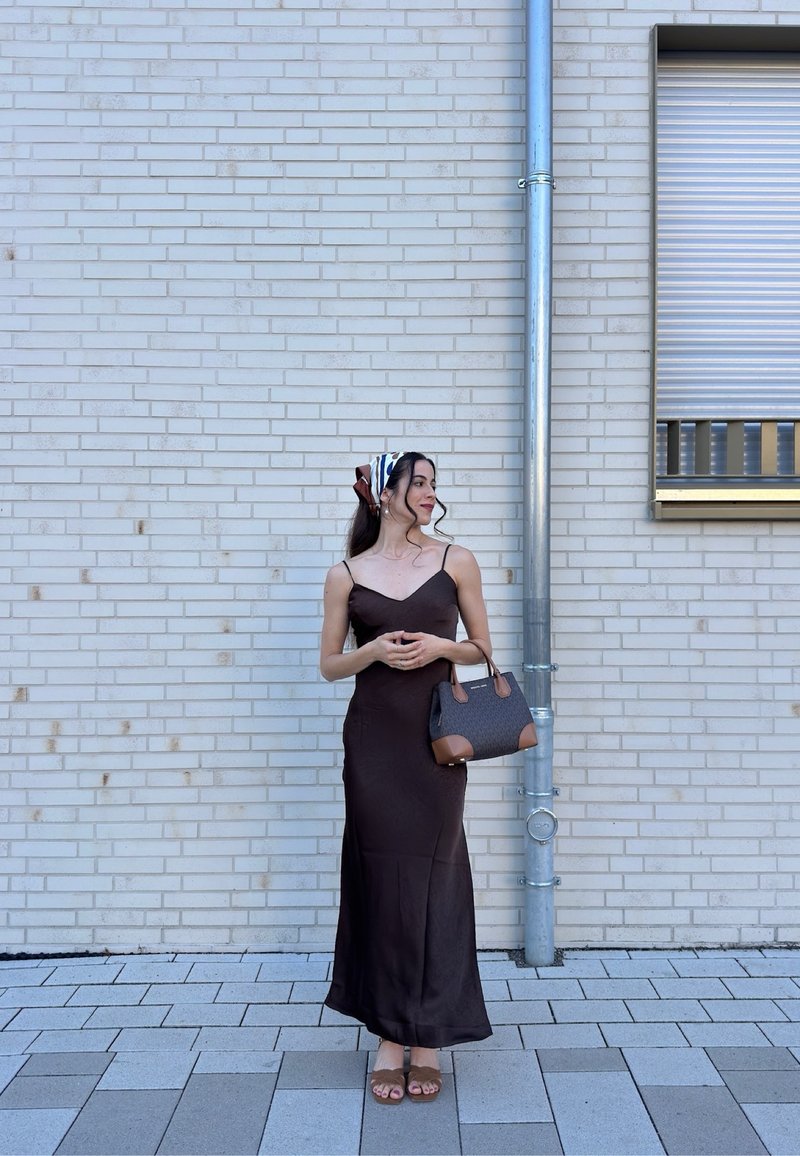 Woman in a long dark dress and headscarf stands on a tiled sidewalk against a light brick wall, holding a handbag and looking sideways.