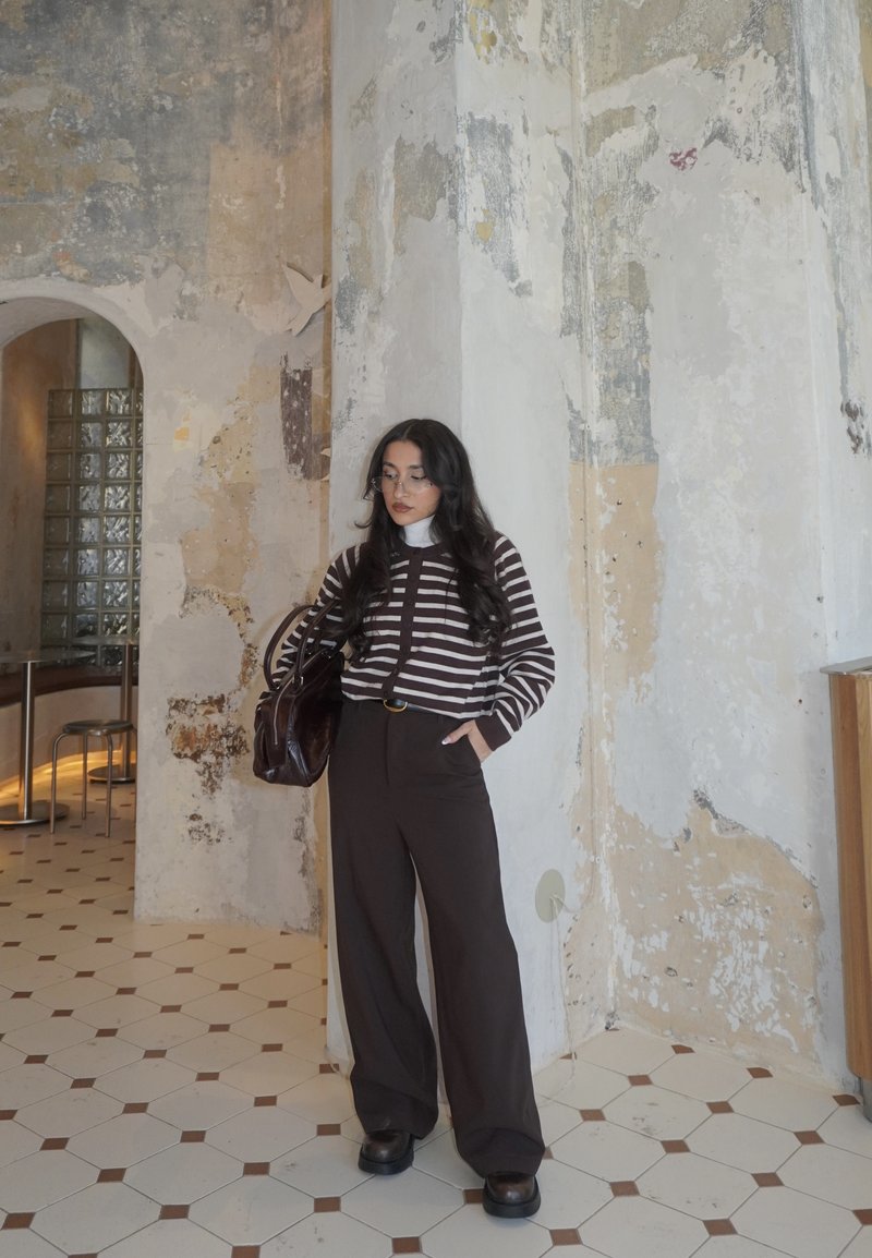 Mujer con cabello largo y oscuro, vestida con un suéter a rayas, pantalones anchos negros y gafas, de pie cerca de una pared texturizada en una habitación con azulejos, sosteniendo una bolsa.