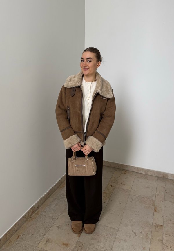 Frau steht drinnen und trägt eine braune Lammfelljacke, schwarze Hose, braune Schuhe und hält eine beige Handtasche.