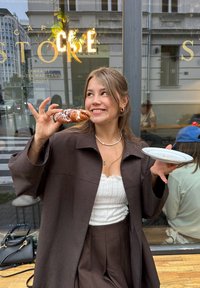 Veste oversize marron, haut texturé blanc et pantalon marron taille haute. Tenant une pâtisserie glacée et une assiette blanche devant la fenêtre d'un café.