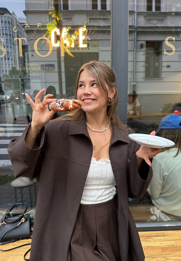 Veste oversize marron, haut texturé blanc et pantalon marron taille haute. Tenant une pâtisserie glacée et une assiette blanche devant la fenêtre d'un café.