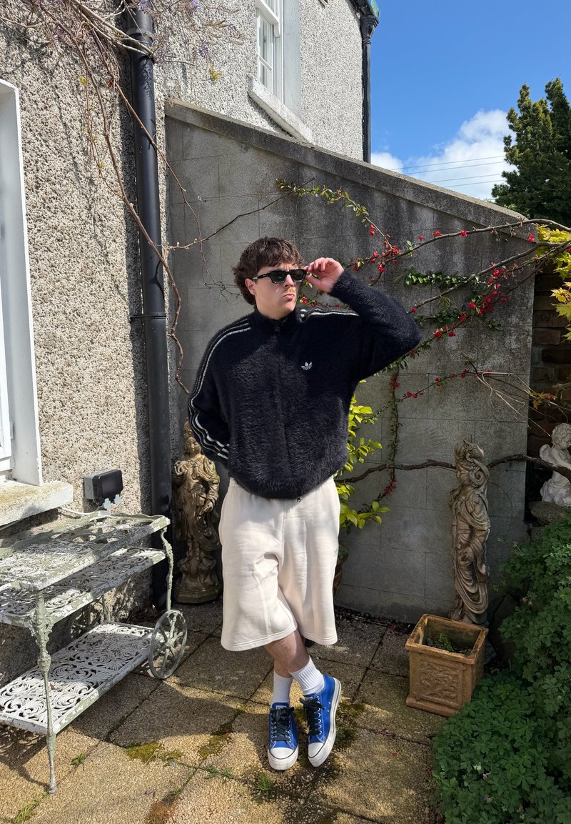 Young man in sunglasses, black fuzzy jacket, white shorts, and blue sneakers stands in sunny garden next to wall with climbing plants and decorative statues.