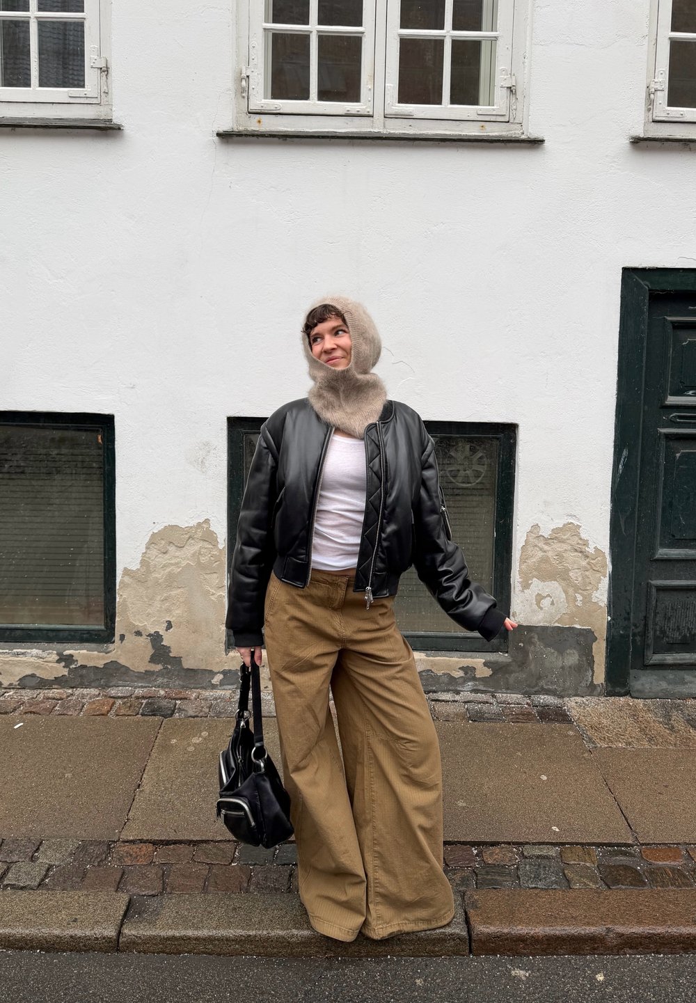 Jeune personne portant une capuche tricotée beige, une veste bomber noire, une chemise blanche, un pantalon large beige clair et un sac à main noir, debout dans une rue pavée.